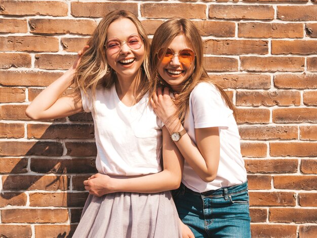 Dos jóvenes hermosas rubias sonrientes chicas hipster en ropa de moda verano camiseta blanca. . Modelos positivos divirtiéndose en gafas de sol