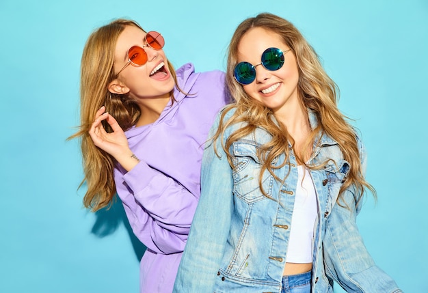 Dos jóvenes hermosas rubias sonrientes mujeres hipster en ropa de moda de verano. Mujeres despreocupadas sexy posando junto a la pared azul en gafas de sol. Modelos positivos volviéndose locos y abrazándose