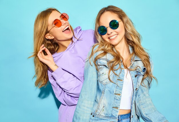 Dos jóvenes hermosas rubias sonrientes mujeres hipster en ropa de moda de verano. Mujeres despreocupadas sexy posando junto a la pared azul en gafas de sol. Modelos positivos volviéndose locos y abrazándose