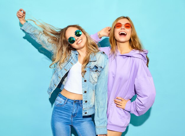 Dos jóvenes hermosas rubias sonrientes mujeres hipster en ropa de moda de verano. Mujeres despreocupadas sexy posando junto a la pared azul en gafas de sol. Modelos positivos volviéndose locos