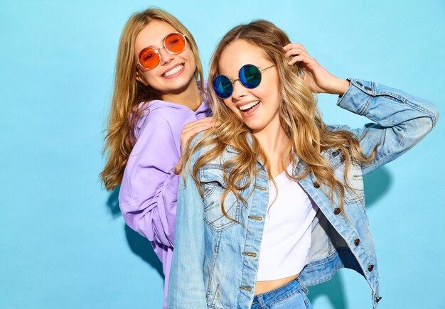 Dos jóvenes hermosas rubias sonrientes mujeres hipster en ropa de moda de verano. Mujeres despreocupadas sexy posando junto a la pared azul en gafas de sol. Modelos positivos volviéndose locos