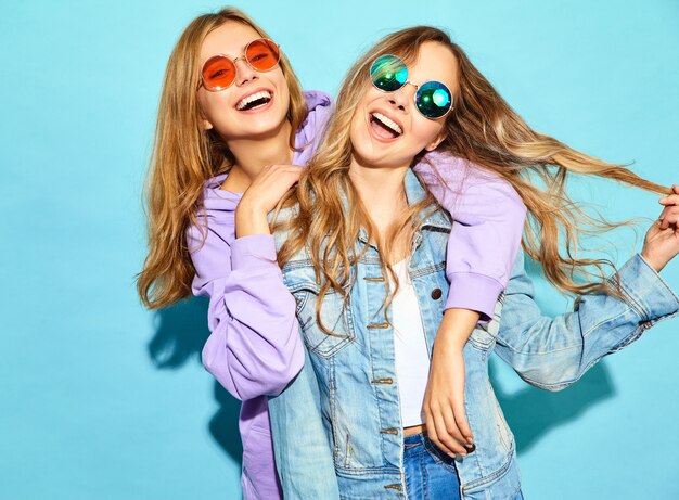 Dos jóvenes hermosas rubias sonrientes mujeres hipster en ropa de moda de verano. Mujeres despreocupadas sexy posando junto a la pared azul en gafas de sol. Modelos positivos