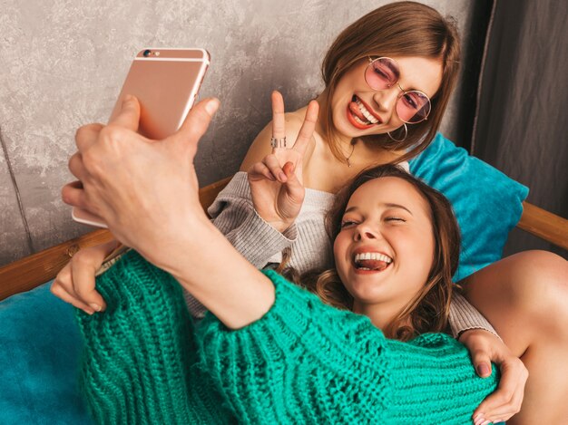 Dos jóvenes hermosas sonrientes hermosas chicas en ropa de moda de verano. Mujeres despreocupadas sexy posando en el interior y tomando selfie. Modelos positivos divirtiéndose con el teléfono inteligente. Mostrando paz