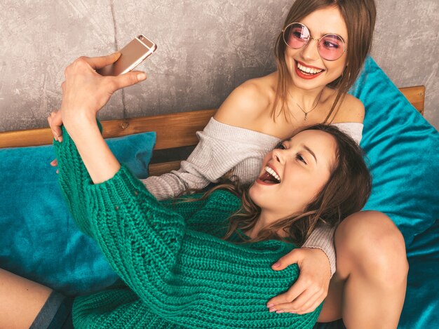 Dos jóvenes hermosas sonrientes hermosas chicas en ropa de moda de verano. Mujeres despreocupadas sexy posando en el interior y tomando selfie. Modelos positivos divirtiéndose con el teléfono inteligente.