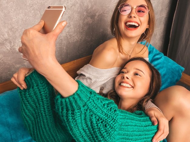 Dos jóvenes hermosas sonrientes hermosas chicas en ropa de moda de verano. Mujeres despreocupadas sexy posando en el interior y tomando selfie. Modelos positivos divirtiéndose con el teléfono inteligente.