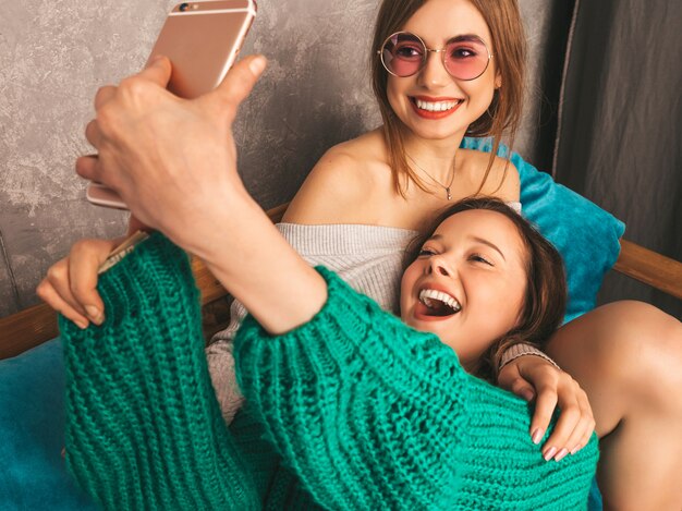 Dos jóvenes hermosas sonrientes hermosas chicas en ropa de moda de verano. Mujeres despreocupadas sexy posando en el interior y tomando selfie. Modelos positivos divirtiéndose con el teléfono inteligente.