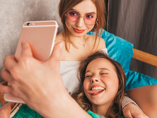 Foto gratuita dos jóvenes hermosas sonrientes hermosas chicas en ropa de moda de verano. mujeres despreocupadas sexy posando en el interior y tomando selfie. modelos positivos divirtiéndose con el teléfono inteligente.