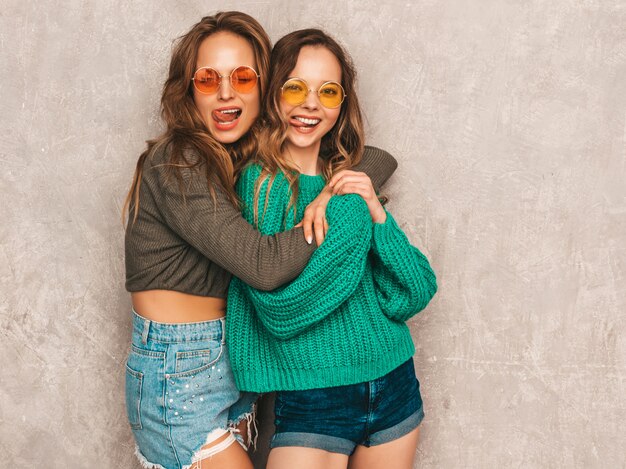 Dos jóvenes hermosas sonrientes hermosas chicas en ropa de moda de verano. Sexy mujer despreocupada posando. Modelos positivos divirtiéndose en gafas de sol redondas. Mostrando la lengua