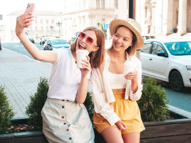 Dos jóvenes hermosas y sonrientes mujeres hipster vestidas de verano. Mujeres sexy y despreocupadas posando en la calle. Modelos puros positivos divirtiéndose al atardecer. Tomando café o té en un vaso de plástico.