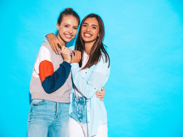 Dos jóvenes hermosas sonrientes rubias hipster chicas en ropa de verano colorida camiseta. Mujeres despreocupadas sexy posando junto a la pared azul. Modelos positivos divirtiéndose