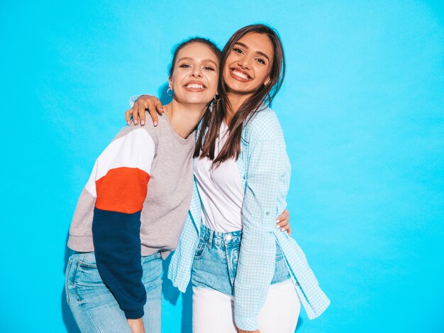 Dos jóvenes hermosas sonrientes rubias hipster chicas en ropa de verano colorida camiseta. Mujeres despreocupadas sexy posando junto a la pared azul. Modelos positivos divirtiéndose