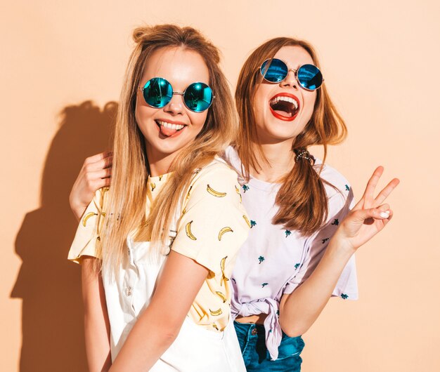 Dos jóvenes hermosas sonrientes rubias hipster chicas en ropa de verano colorida camiseta. Mujeres despreocupadas sexy posando junto a la pared de color beige en gafas de sol redondas. Mostrando signo de paz