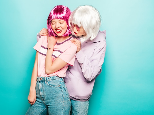 Dos jóvenes muchachas sonrientes sexy hipster en pelucas y labios rojos. Hermosas mujeres de moda en ropa de verano. Modelos despreocupados posando junto a la pared azul en el estudio volviéndose loco y abrazándose