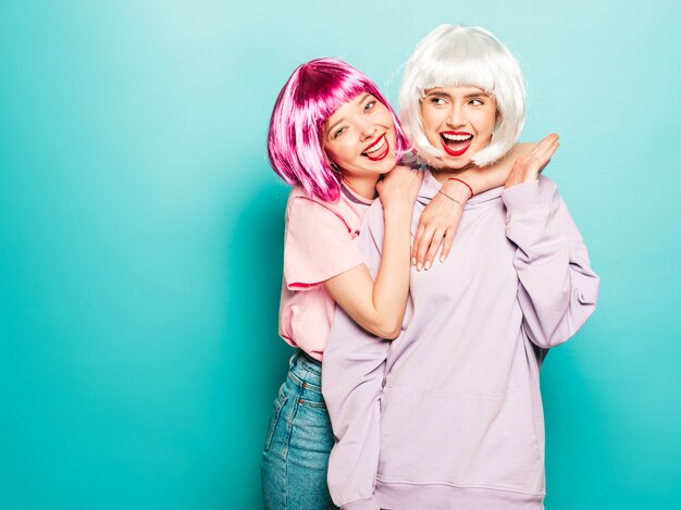 Dos jóvenes muchachas sonrientes sexy hipster en pelucas y labios rojos. Hermosas mujeres de moda en ropa de verano. Modelos despreocupados posando junto a la pared azul en el estudio volviéndose loco y abrazándose