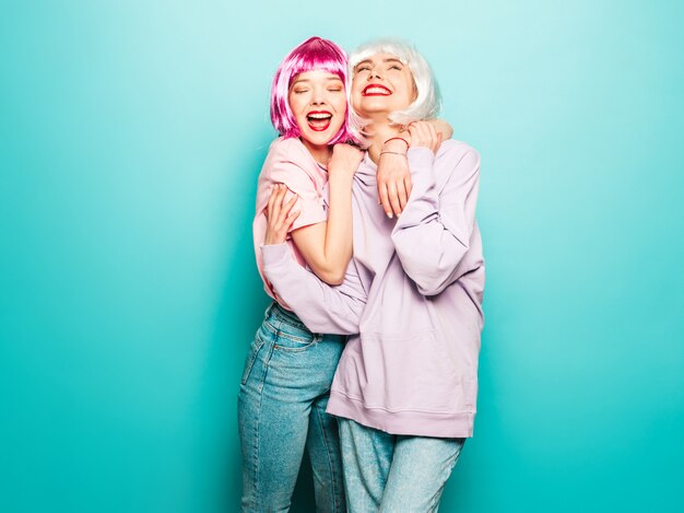 Dos jóvenes muchachas sonrientes sexy hipster en pelucas y labios rojos. Hermosas mujeres de moda en ropa de verano. Modelos despreocupados posando junto a la pared azul en el estudio volviéndose loco y abrazándose