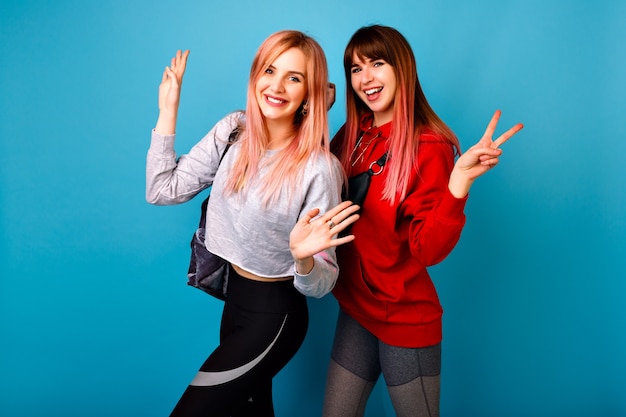 Dos jóvenes mujeres hipster bonitas divertidas con atuendos casuales brillantes deportivos, sonriendo gritando y saludando, pared azul