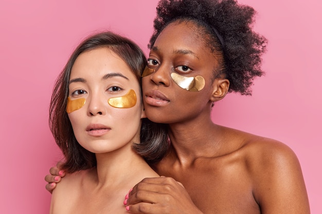 Dos jóvenes mujeres multiétnicas se aplican parches dorados debajo de los ojos, de pie cerca uno del otro, tienen una piel suave, limpia y saludable, disfrutan del día de spa y belleza aislado en la pared rosa