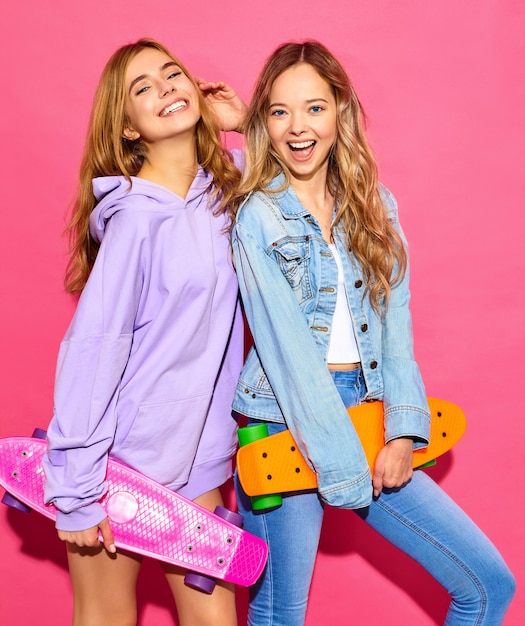 Dos jóvenes mujeres rubias sonrientes con estilo con patinetas centavo. Modelos en ropa deportiva de verano hipster posando junto a la pared de color rosa. Mujeres positivas volviéndose locas