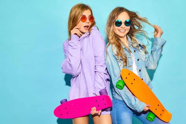 Foto gratuita dos jóvenes mujeres rubias sonrientes con estilo con patinetas centavo. mujeres en ropa deportiva de verano hipster posando junto a la pared azul. modelos positivos