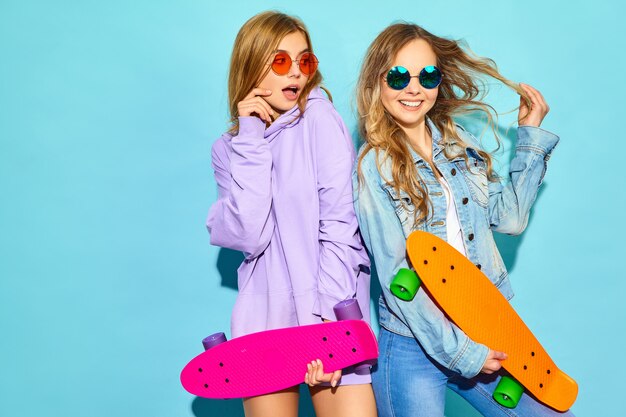 Dos jóvenes mujeres rubias sonrientes con estilo con patinetas centavo. Mujeres en ropa deportiva de verano hipster posando junto a la pared azul. Modelos positivos