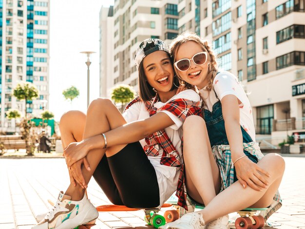 Dos jóvenes sonrientes hermosas chicas con patinetas coloridas centavo. Mujeres en ropa hipster de verano sentado en el fondo de la calle. Modelos positivos divirtiéndose y volviéndose locos. Mostrando lenguas
