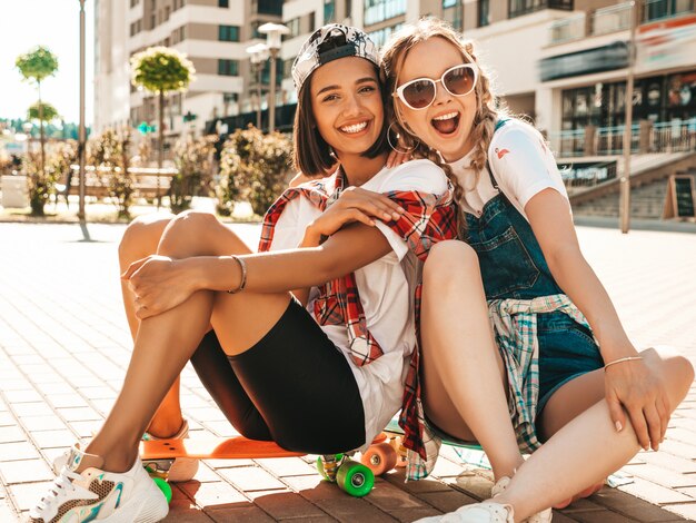 Dos jóvenes sonrientes hermosas chicas con patinetas coloridas centavo. Mujeres en ropa hipster de verano sentado en el fondo de la calle. Modelos positivos divirtiéndose y volviéndose locos