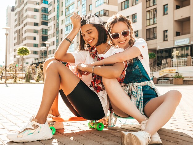 Dos jóvenes sonrientes hermosas chicas con patinetas coloridas centavo. Mujeres en ropa hipster de verano sentado en el fondo de la calle. Modelos positivos divirtiéndose y volviéndose locos