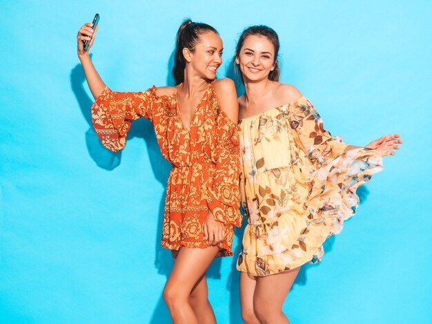 Dos jóvenes sonrientes mujeres hipster en vestidos hippie de verano. Chicas tomando fotos de autorretrato en smartphone. Modelos posando junto a la pared azul en el estudio