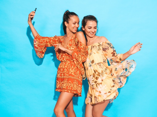 Dos jóvenes sonrientes mujeres hipster en vestidos de verano hippie volando. Chicas tomando fotos de autorretrato en smartphone. Modelos posando junto a la pared azul en el estudio. Mujer mostrando emociones positivas.