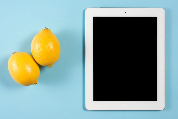Dos jugosos limones amarillos cerca de la tableta digital con pantalla negra sobre fondo azul