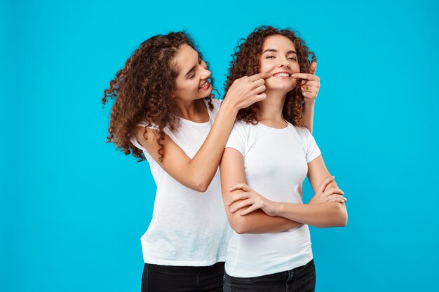 Dos lindas gemelas sonrientes, bromeando sobre la pared azul