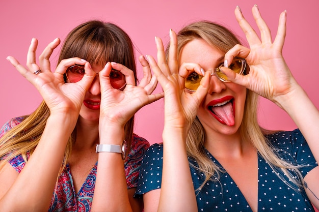 Foto gratuita dos locas mujer divertida imitando vasos con sus manos, mejor amigo de fiesta, pared rosa, vestidos elegantes, signos de gestos.