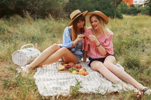 Dos maravillosas chicas con sombrero de paja pasando las vacaciones en el campo, bebiendo vino espumoso.