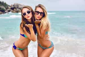 Foto gratuita dos mejores amigas jóvenes morenas y rubias que te envían un beso al aire, tienen un cuerpo delgado y sexy, usan gafas de sol de bikini y joyas brillantes de moda, posando frente a una playa tropical.