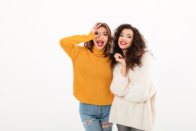 Dos muchachas alegres en suéteres divirtiéndose juntos sobre la pared blanca