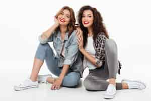 Foto gratuita dos muchachas sonrientes que se sientan juntas en el piso sobre la pared blanca