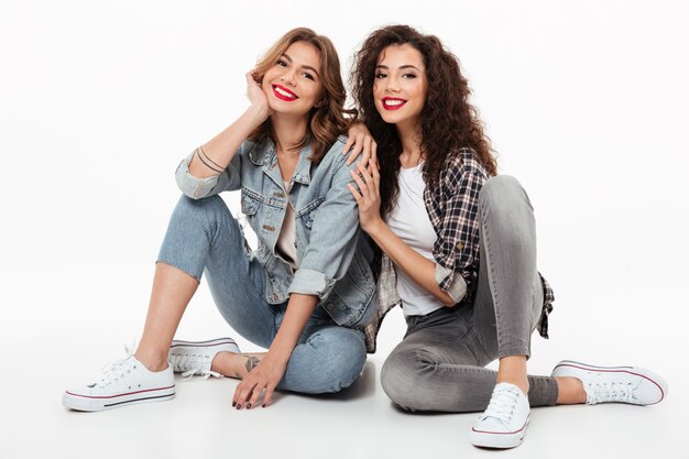 Dos muchachas sonrientes que se sientan juntas en el piso sobre la pared blanca