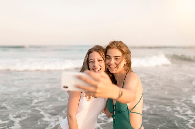Dos muchachas sonrientes que toman el autorretrato del teléfono celular cerca de la costa