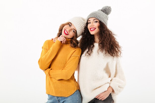 Dos muchachas sonrientes en suéteres y sombreros posando juntos sobre pared blanca