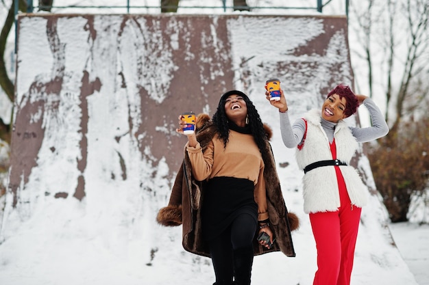 Dos mujeres afroamericanas con piel de oveja y abrigo de piel posaron en el día de invierno contra un fondo nevado con tazas de café