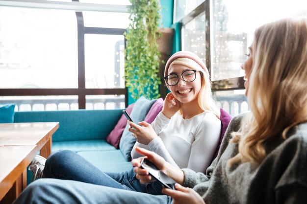 Dos mujeres alegres usando teléfonos celulares y riendo en café