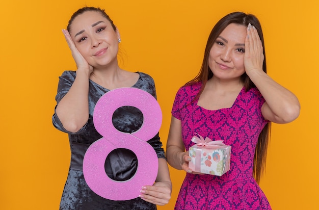 Dos mujeres asiáticas celebrando el día internacional de la mujer sosteniendo el número ocho y presente mirando al frente confundidos tomados de la mano en la cabeza de pie sobre la pared naranja