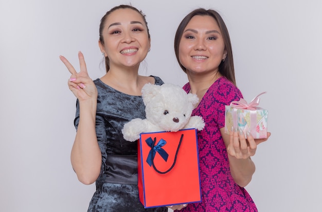 Foto gratuita dos mujeres asiáticas felices que sostienen una bolsa de papel con un oso de peluche y un presente celebrando el día internacional de la mujer sonriendo alegremente mostrando el signo v de pie sobre la pared blanca