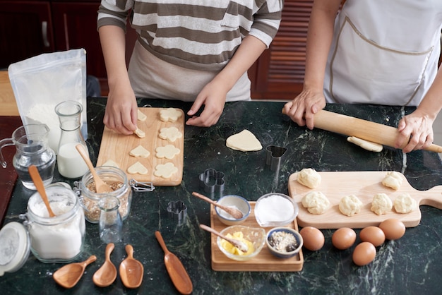 Dos mujeres asiáticas irreconocibles enrollando masa y cortando galletas en la encimera de la cocina