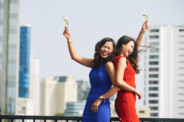 Dos mujeres asiáticas con vestidos brillantes sosteniendo copas de champán en una fiesta urbana en la azotea