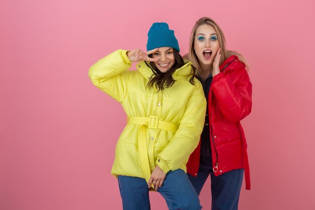 Dos mujeres atractivas amigas tomando fotos selfie en pared rosa en colorida chaqueta de invierno de color rojo y amarillo brillante divirtiéndose juntos, tendencia de moda de ropa deportiva de abrigo cálido, loco divertido