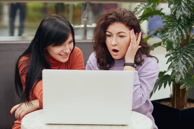 Dos mujeres atractivas con una computadora portátil blanca sentada en caffe, mirando la pantalla del portátil con expresiones faciales asombradas, ven algo interesante mientras descansan en la cafetería.