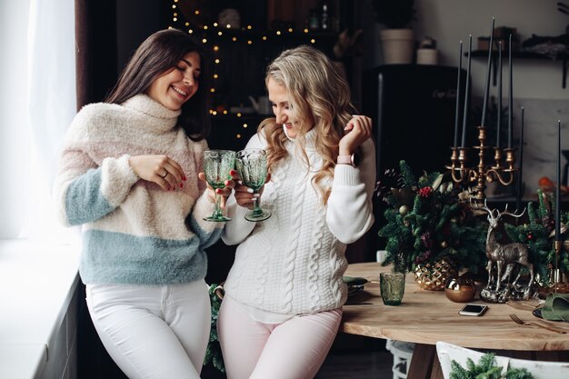 Dos mujeres atractivas en suéteres brindando vasos de bebidas en la mesa de la cocina con adornos navideños.