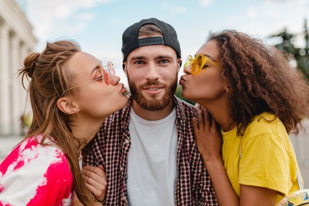 Dos mujeres besando al hombre, feliz emoción emocionada, joven compañía de amigos sonrientes divirtiéndose juntos