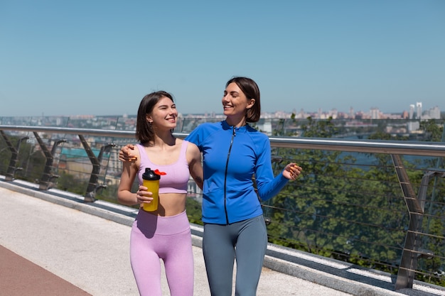 Dos mujeres bonitas en ropa deportiva en amigos del puente hablan felices y positivas mientras caminan sonríen, disfrutan de la mañana de fitness, increíbles vistas de la ciudad en el fondo
